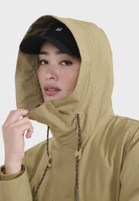 Veste imperméable beige avec un col haut et une capuche ajustable. Elle comprend des cordons de serrage et des boutons, associée à une casquette noire.