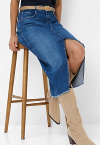 Denim midi-rok met een split aan de voorkant, gefranjerde zoom, blauwe stof en zakken. Gecombineerd met beige knie-lange laarzen met decoratieve stiksels.