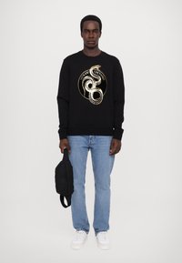 Felpa nera con grafiche dorate di un serpente, indossata con jeans azzurri chiari e sneakers bianche, accompagnata da una borsa nera.
