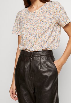 Femme portant un chemisier blanc à manches courtes avec un petit motif floral orange, bleu et jaune, rentré dans un pantalon en cuir marron foncé à taille haute.