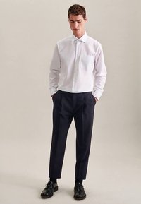 Witte overhemd met lange mouwen, een button-down kraag en een borstzak, gecombineerd met donkerblauwe broek en zwarte schoenen, tegen een neutrale achtergrond.