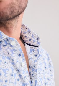 Detailný záber na mužský krk a bradu, zobrazujúci golier svetlomodrej kvetinovej košele s bielymi gombíkmi a tmavým lemom na okraji.