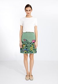 Haut blanc à manches courtes associé à une jupe verte ornée de broderies florales colorées, avec des accents bleus, jaunes et roses. Texture lisse.