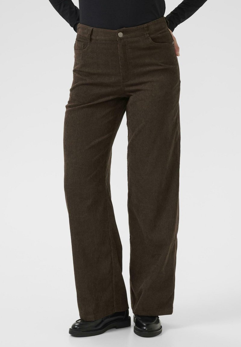 Personne portant un pantalon large en velours côtelé marron foncé et des chaussures noires, debout, les mains sur les hanches, sur un fond uni.