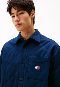 Jaunas vyras dėvi tamsiai mėlyną siuvinėtą striukę su priekiniais krūtinės kišenėmis, ant kurių yra Tommy Jeans logotipo pagalvėlė.