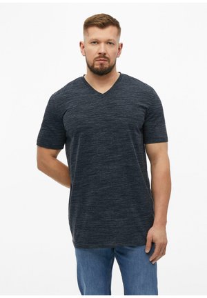 Mężczyzna z krótkimi włosami i brodą, ubrany w ciemnoszary T-shirt z krótkim rękawem i dekoltem w serek oraz niebieskie dżinsy, stojący na jednolitym białym tle.