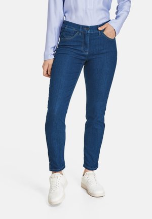 Frau trägt blaue Slim-Fit-Jeans, hellblaue Bluse und weiße Sneakers, steht mit einer Hand in der Tasche.
