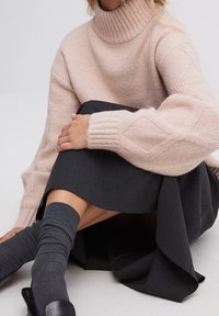 En person sitter med benen i kors iförd en blek rosa polotröja, mörkgrå kjol, kolgrå stickade strumpor och svarta skor.