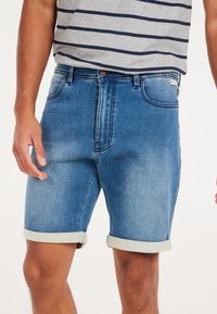 Bermudas de mezclilla en un degradado de azul claro a oscuro, con un dobladillo enrollado, cinco bolsillos y cierre de botón. Combinadas con una camisa de rayas gris.