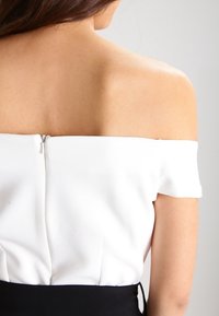 Femme aux longs cheveux bruns portant un haut blanc sans épaules avec une fermeture éclair dans le dos, montrant ses épaules nues et le haut de son dos.
