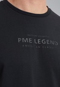 Crna pamučna majica s okruglim izrezom i urezanim tekstom: "NEOGRANIČENA SLOBODA PME LEGENDA AMERIČKA KLASIKA" na prednjoj strani.