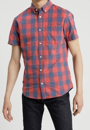 Mężczyzna w koszuli z krótkim rękawem zapinanej na guziki z czerwono-niebieskim wzorem w kratę i czarnych spodniach, stojący na jednolitym tle.