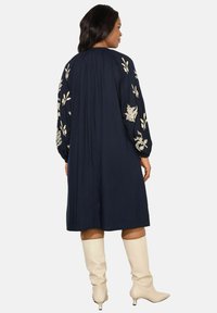 Marineblauwe jurk met lange, geborduurde mouwen met bloemenpatronen. De jurk is tot op de knie, gecombineerd met romige laarzen tot de knie.