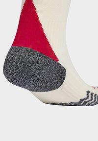 Detailní záběr na oblast paty a kotníku ponožky s červenými, bílými a černými strukturovanými pletenými vzory na jednolitém pozadí.