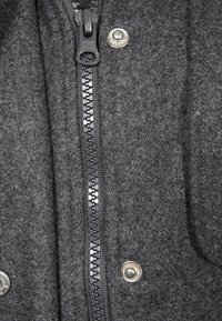 Grå uldjakke med en tekstureret finish, der har en sort lynlås midt på og sølvfarvede trykknapper på hver side.