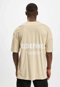 Beige bomull t-skjorte med oversized passform, korte ermer og fet hvit tekst på ryggen, inkludert "E(DEFINED)" og merkevaren logo.