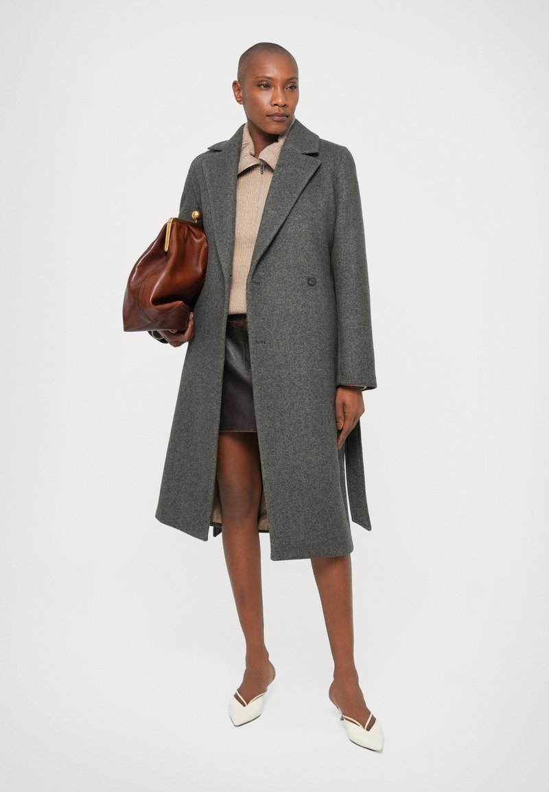 Grauer Wollmantel mit doppelreihigem Schnitt, Reverskragen und Seitentaschen. Kombiniert mit einem beigen Strickoberteil und einer braunen ledernen Handtasche.