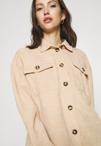 Béžová kordurová košele-jacket s button-down prednou stranou, dvoma náprsnými vreckami a kontrastnými hnedými gombíkmi. Mäkká textúra a uvoľnený strih.