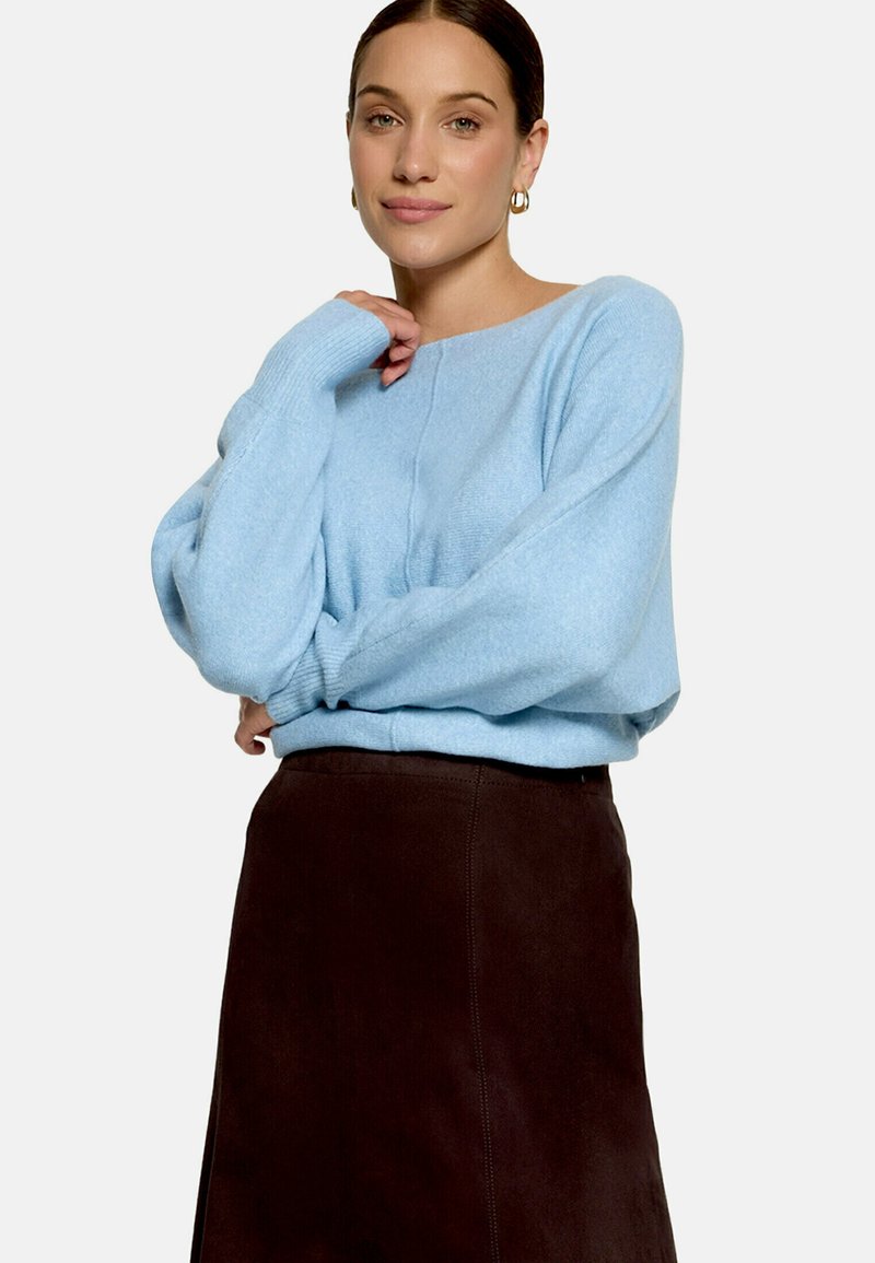 Femme portant un pull bleu clair et une jupe marron foncé, posant avec une main près de son visage sur un fond blanc uni.