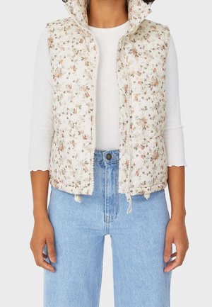 Mujer vistiendo un chaleco sin mangas con un estampado floral sobre una blusa blanca, combinado con unos vaqueros de tiro alto de color azul claro.
