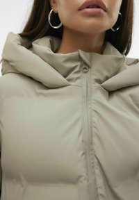 Veste à capuche rembourrée de couleur beige clair, dotée d'une fermeture éclair et d'une texture de tissu douce et lisse. Capuche proéminente avec des détails froncés.