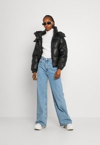 Mujer con cabello trenzado que lleva gafas de sol negras, una chaqueta acolchada negra, un cuello de tortuga blanco, unos vaqueros de pierna ancha de color azul claro y zapatillas blancas.