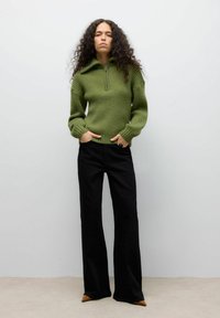 Maglione verde lavorato a maglia con collo a zip, costine testurizzate e maniche ampie, abbinato a pantaloni neri a zampa d'elefante e tacchi a punta marroni.