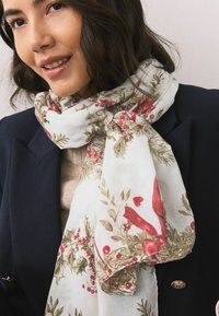 Lehký průsvitný šátek s květinovým a ptáčím potiskem v červené a zelené barvě na bílém pozadí, volně omotaný kolem krku.