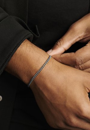Silberfarbener Armreif mit geflochtenem Design, am Handgelenk getragen. Hautfarbe sichtbar unter dem Armreif, kontrastiert mit dunkler Kleidung.