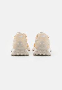Un paio di sneakers New Balance beige e arancioni viste da dietro, con suole del tallone triangolari e texturizzate, su uno sfondo bianco.