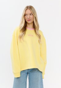 Żółta, oversize'owa bluza z różowym wykończeniem kołnierzyka, z małym haftowanym logo z przodu. Nosi się ją z jasnoniebieskimi jeansami.