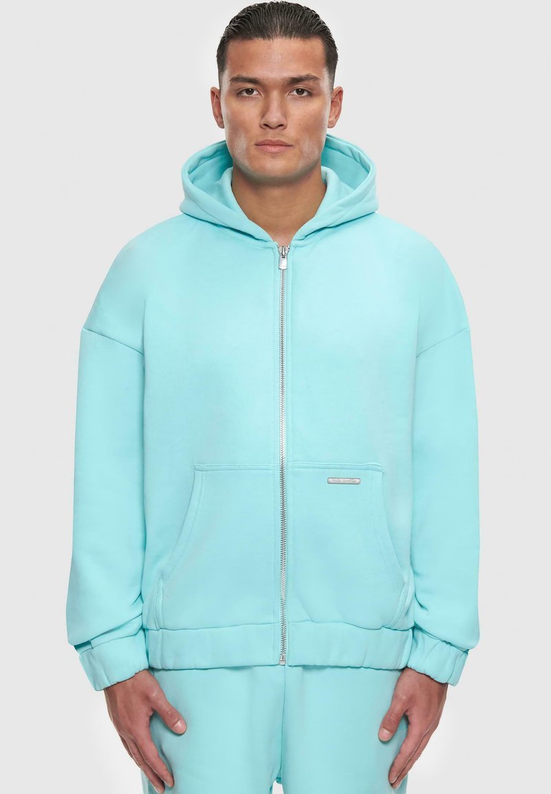 Felpa con zip turchese realizzata in tessuto morbido. Caratterizzata da un ampio cappuccio, tasche frontali e polsini a coste. Design semplice con hardware minimo.