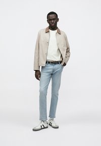 Giacca beige chiaro con colletto marrone, felpa bianca, jeans azzurri e sneakers bianche con strisce nere. Look semplice e casual.