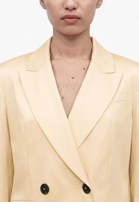 Άτομο που φοράει ανοιχτό κίτρινο σακάκι διπλής σειράς με μαύρα κουμπιά, εμφανίζοντας το πάνω μέρος του στήθους και τον αυχένα πάνω σε απλό φόντο.