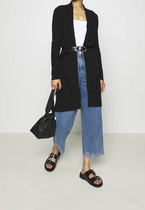 Larga chaqueta negra con un frente abierto, combinada con vaqueros azul de tiro alto con un dobladillo floral con volantes y sandalias negras sin talón.