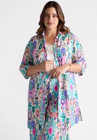 Camicia leggera con bottoni in un motivo floreale multicolore che presenta tonalità di verde, blu, rosa e viola; maniche lunghe arrotolate sui gomiti.