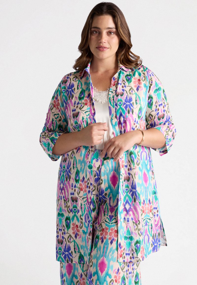 Camicia leggera con bottoni in un motivo floreale multicolore che presenta tonalità di verde, blu, rosa e viola; maniche lunghe arrotolate sui gomiti.