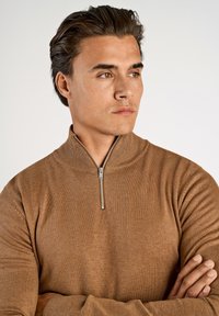 Brauner gerippter Strickpullover mit hohem Kragen und halbem Reißverschluss, der eine weiche Textur und eine figurbetonte Passform aufweist.
