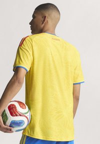 Gul sportsjersey med et struktureret mønster, korte ærmer, blå detaljering ved manchetten og røde striber på skuldrene. Spilleren holder en multicolor fodbold.