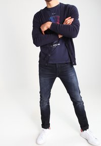 Cárdigan navy con botones, superpuesto a una camiseta oscura con un diseño geométrico, combinado con vaqueros oscuros y zapatillas blancas.