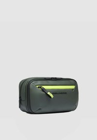 Donkergroene reistas gemaakt van glad, duurzaam materiaal. Voorzien van een zwarte ritssluiting met een felgroene accentkleur en een logo aan de voorkant. Rechthoekige vorm.