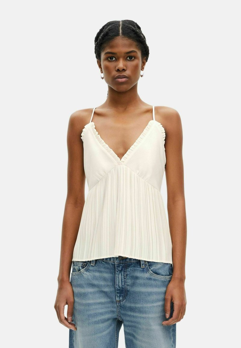 Witte geplooide top met ruffles aan de halslijn en dunne verstelbare bandjes, gedragen met lichtblauwe versleten denimjeans.