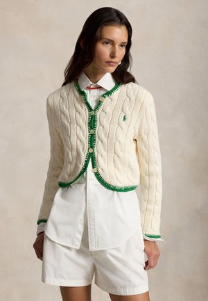 Nő krémszínű, zöld szegélyű, kötött kardigánt visel fehér gombos ing és fehér rövidnadrág felett, semleges arckifejezéssel áll.