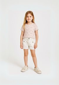 Vaaleanpunainen lyhythihainen paita, jossa on pieniä kukkakuviota, yhdistettynä valkoisiinShortseihin, joissa on napitusyksityiskohtia. Mallilla on päällään beigeet lenkkarit.