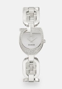 Montre en argent avec un cadran circulaire, ornée de cristaux transparents, bracelet en métal maillé, et repères horaires minimalistes. Le nom de la marque est visible.