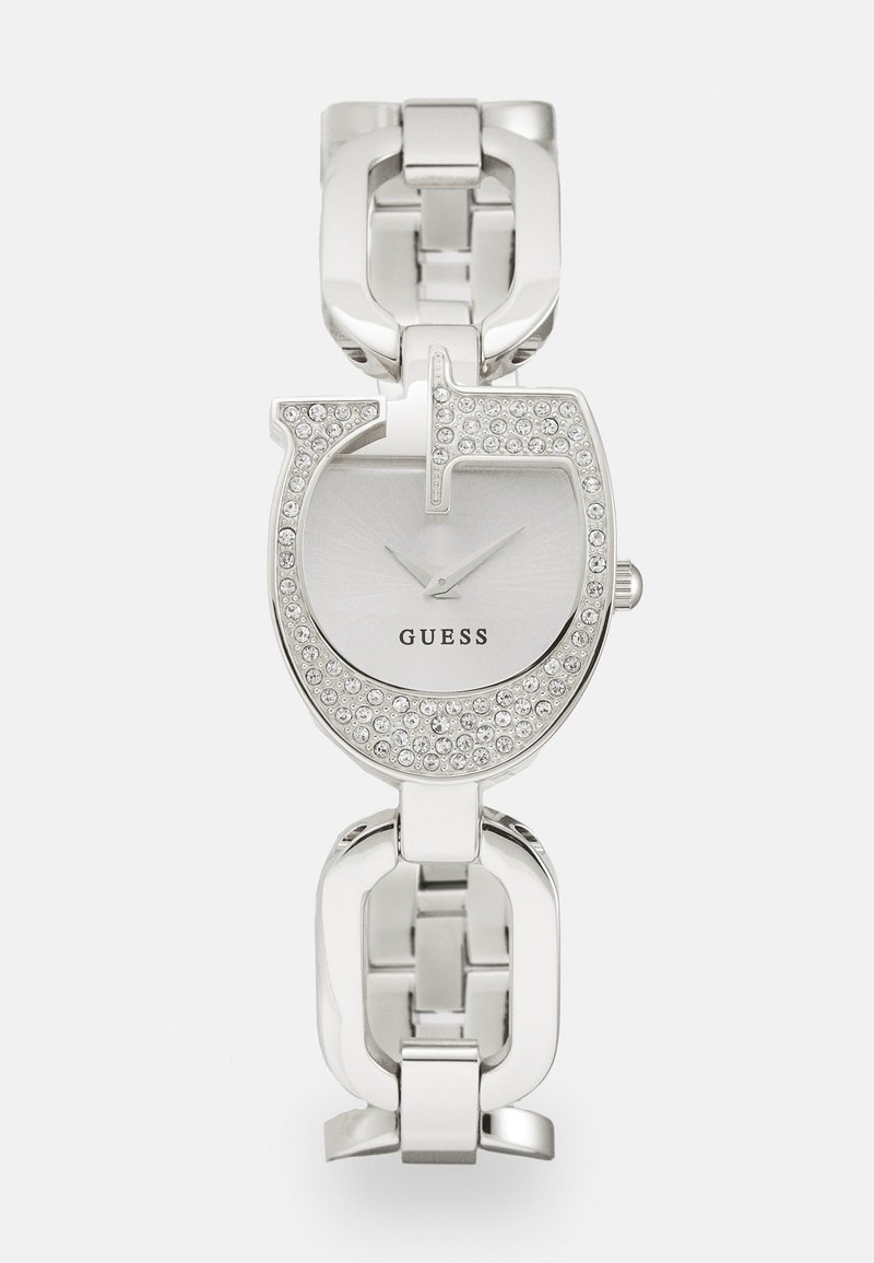 Montre en argent avec un cadran circulaire, ornée de cristaux transparents, bracelet en métal maillé, et repères horaires minimalistes. Le nom de la marque est visible.
