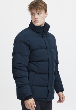 Giacca imbottita blu navy con colletto alto, tasche laterali e polsini elasticizzati. Tessuto liscio e strutturato con chiusura a zip.