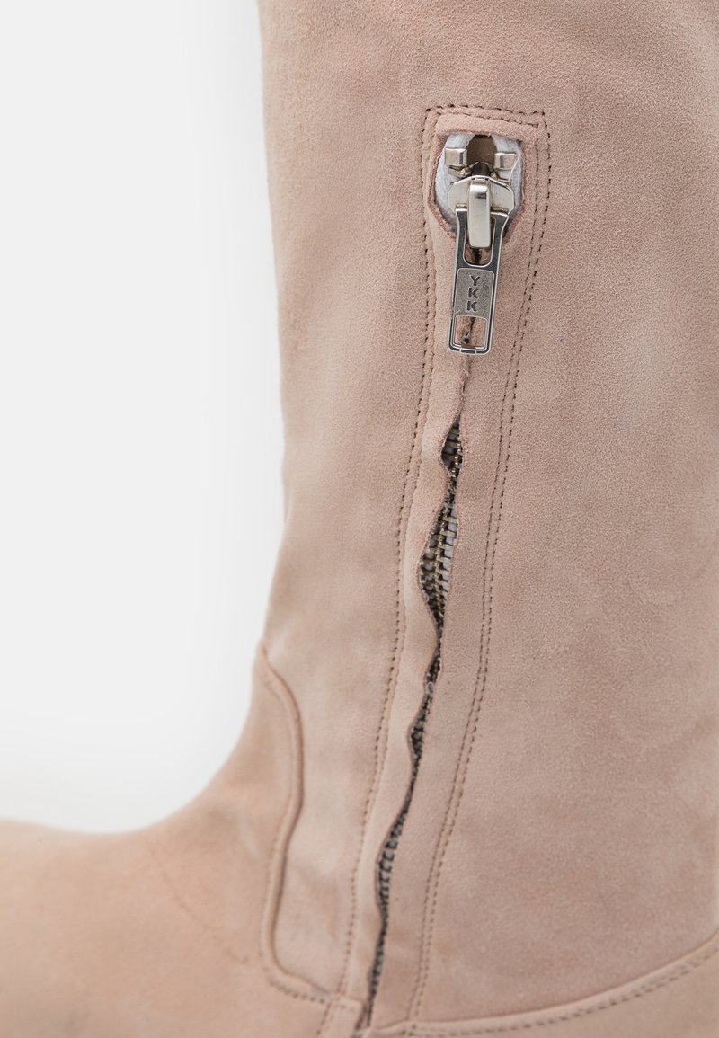 Beige Wildleder kniehohe Stiefel mit einem seitlichen YKK-Reißverschluss und einer glatten Textur mit genähten Details entlang der Nähte.