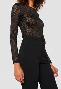 Svart spetsbodysuit med långa ärmar och blommönster, kombinerad med figurnära svarta byxor. Slätt tyg och elegant silhuett.