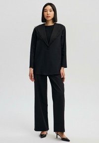 Černý blazer s texturovaným límcem, zapínání na jeden knoflík, kombinovaný s širokými černými kalhotami a špičatými černými podpatky. Hladký povrch látky.